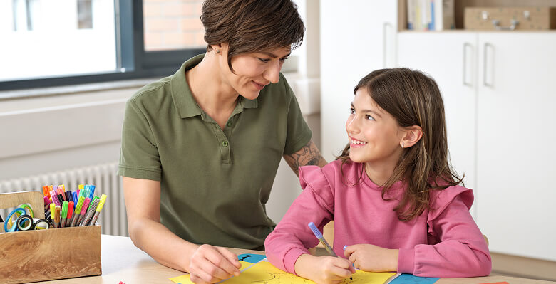 Senior schreibt eine Übung mit Unterstützung einer Betreuerin am Tisch